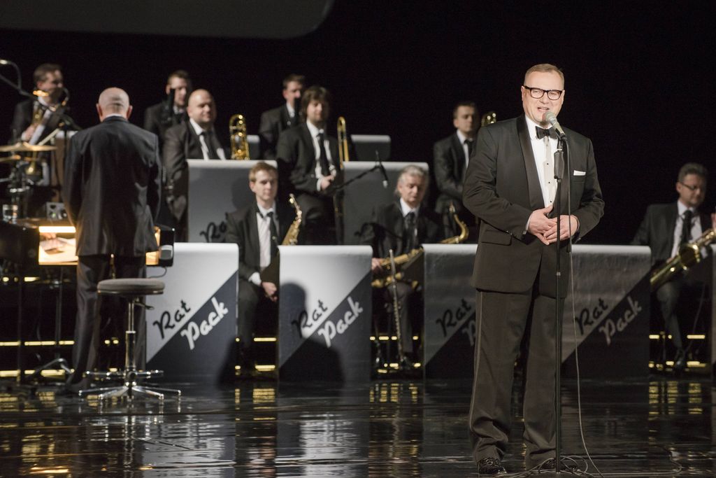 Przy mikrofonie stoi Konferansjer, blondyn w okularach i czarnym smokingu. Za nim widać dyrygenta i muzyków big-bandu, siedzących za pulpitami z napisem „Rat Pack”.