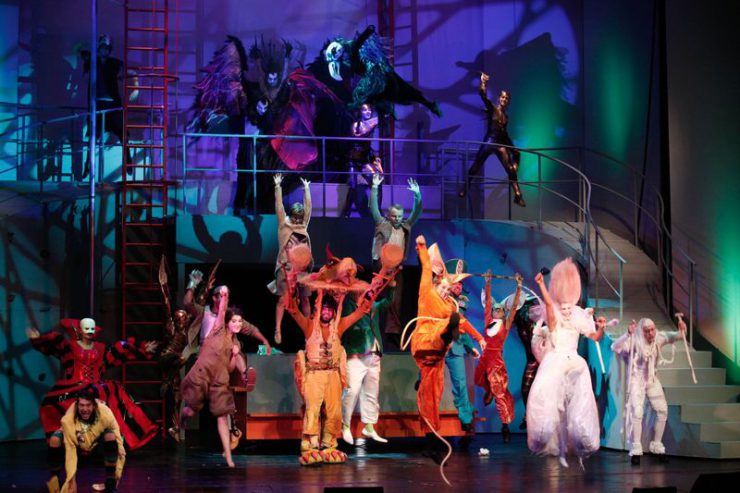 Scena zbiorowa z wysoką konstrukcją ze schodami w tle. Z przodu jasno oświetleni śpiewający i tańczący główni bohaterowie, wyżej na platformie kilka ciemnych postaci.