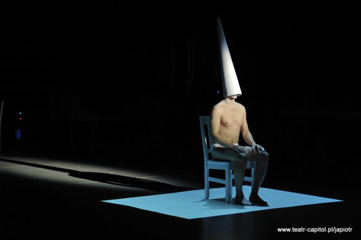 Ciemny kadr z wiązką światła skierowaną na półnagiego człowieka siedzącego na krześle, opierającego dłonie na kolanach. Mężczyzna ma na głowie czapkę w kształcie bardzo wysokiego stożka, zakrywającą mu twarz.