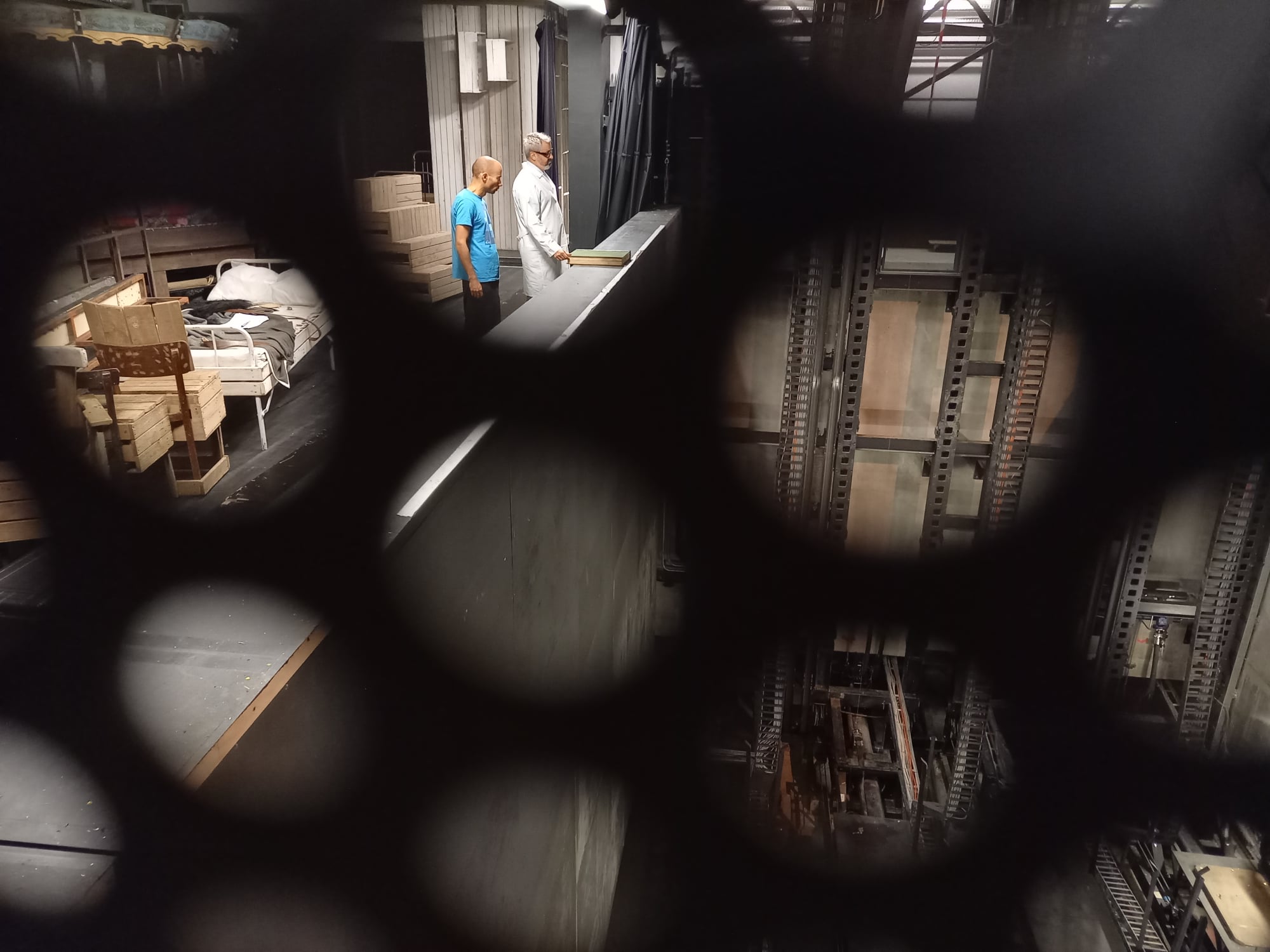 Przez okrągłe otwory ażurowej kraty widać jedną ze scen na planie serialu: aktorzy stoją nad głębokim otworem zapadni, mieszczącej hydrauliczny mechanizm podnoszenia podłogi scenicznej.