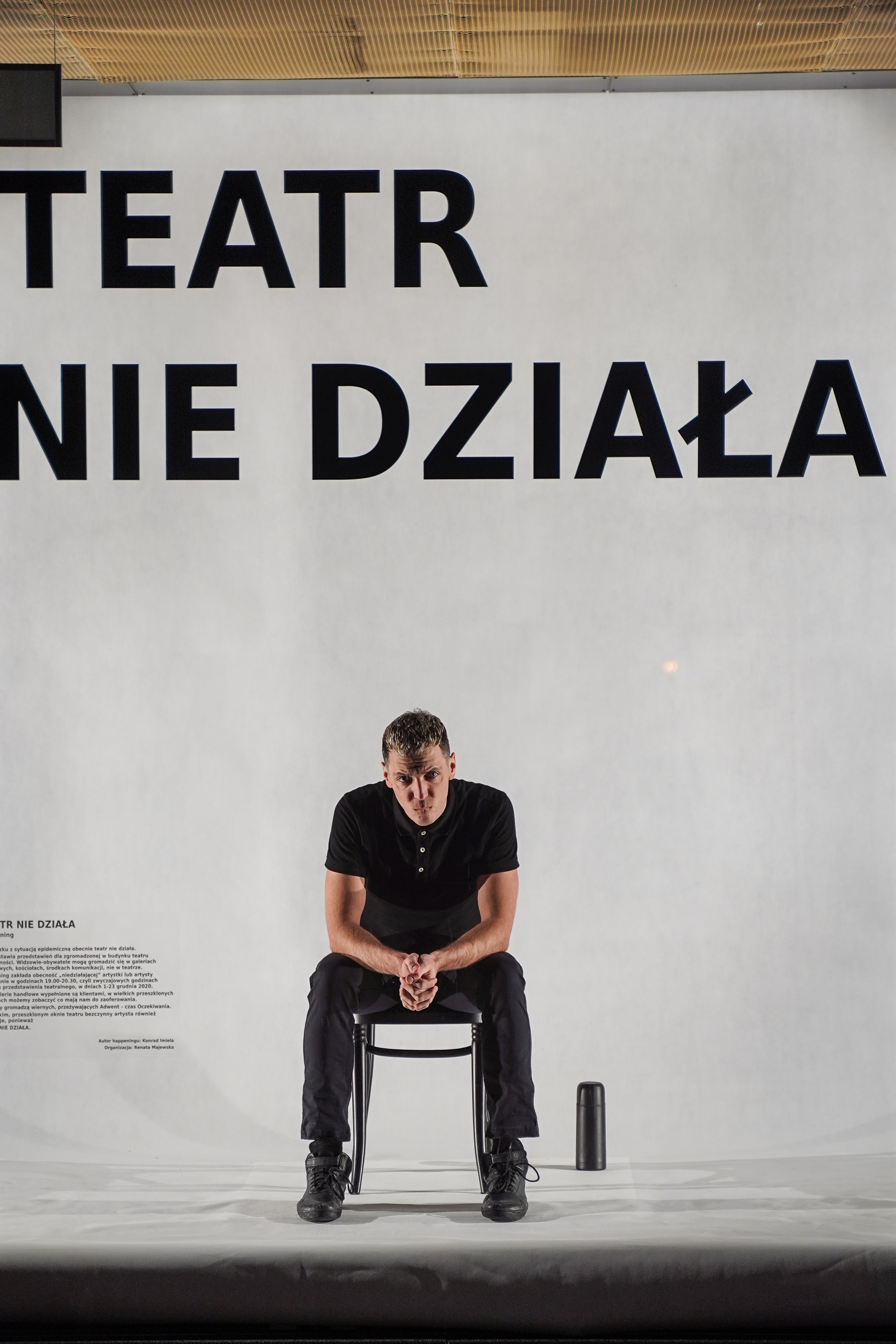 W witrynie teatru, na białym tle, na czarnym krześle siedzi aktor. Na szybie widac duży czarny napis TEATR NIE DZIAŁA
