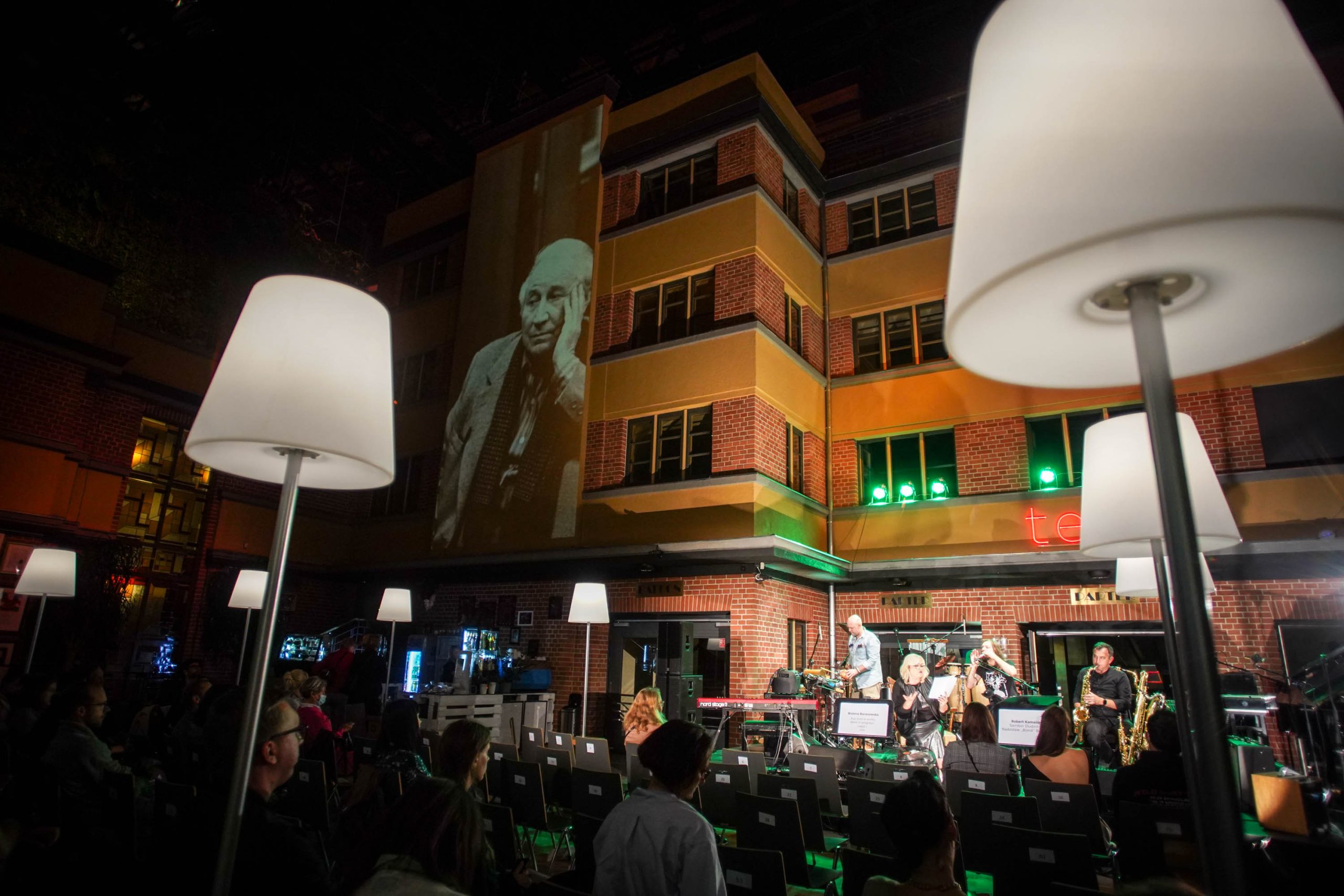 Noc, Podwórko Teatru Muzycznego Capitol. Na tle ceglanej ściany z oknami, wysokiej na trzy piętra, mała jasno oświetlona scena, pełna instrumentów muzycznych. Stoi na niej aktor przed mikrofonem. Ogląda go sopra grupa publiczności, siedzącej na krzesłach miedzy stojącymi lampami o białych kloszach. Na gładkim fragmencie ściany, o wysokości kilkunastu metrów, wyświetlone jest czarno-białe zdjęcie portet Tadeusza Różewicza.