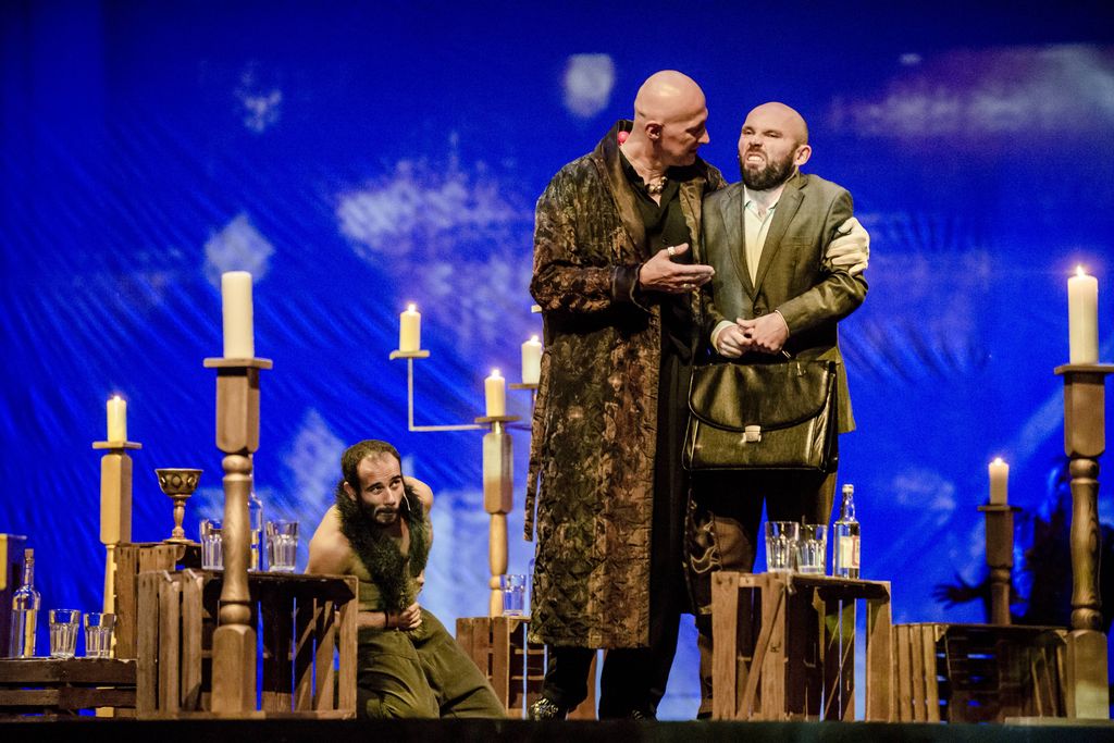 Wśród drewnianych skrzynek, na których i obok których, znajdują się lichtarze i świece, stoi dwóch mężczyzn na pierwszym planie, a jedna postać – Behemot, klęczy z tyłu. Wysoki, łysy Woland w eleganckim, długim, brązowym szlafroku lewą ręką obejmuje człowieka w garniturze i z teczką w dłoniach. Woland coś mówi, głowę ma zwróconą w stronę mężczyzny, kieruje w jego stronę prawą dłoń. Mężczyzna z grymasem na twarzy wygląda na niezadowolonego. Przed nimi, na skrzynce stoi butelka wódki i dwie szklanki.