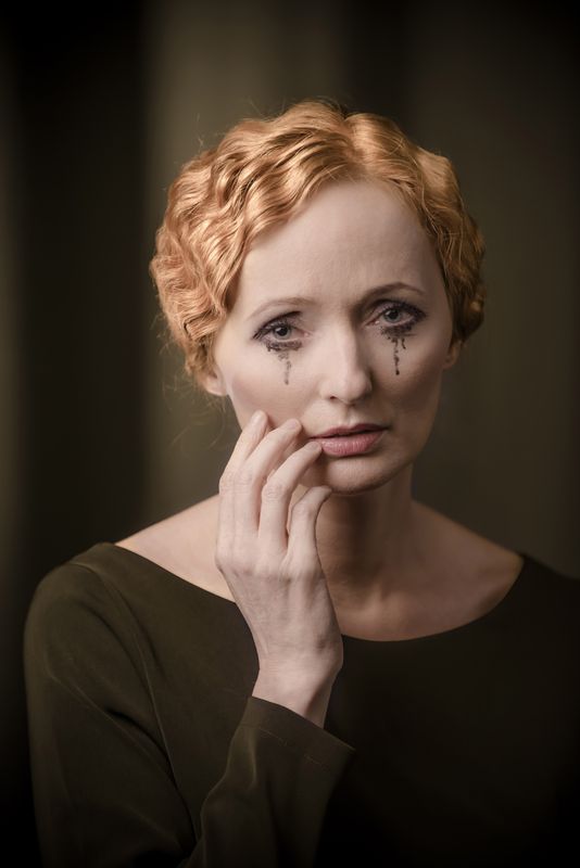 Portret Justyny Szafran kreującej rolę Kobiety. Ciemny strój kontrastuje z bladą twarzą, nieskazitelną cerę podkreśla delikatny makijaż, jedynym zgrzytem jest rozmazany tusz płynący z oczu. Artystka prawą dłonią lekko dotyka prawego policzka i okolicy ust, patrzy ze smutkiem. Ma rude, falujące włosy bez grzywki, spięte z tyłu głowy. Kolorystyka ciemnej zieleni i sepii.